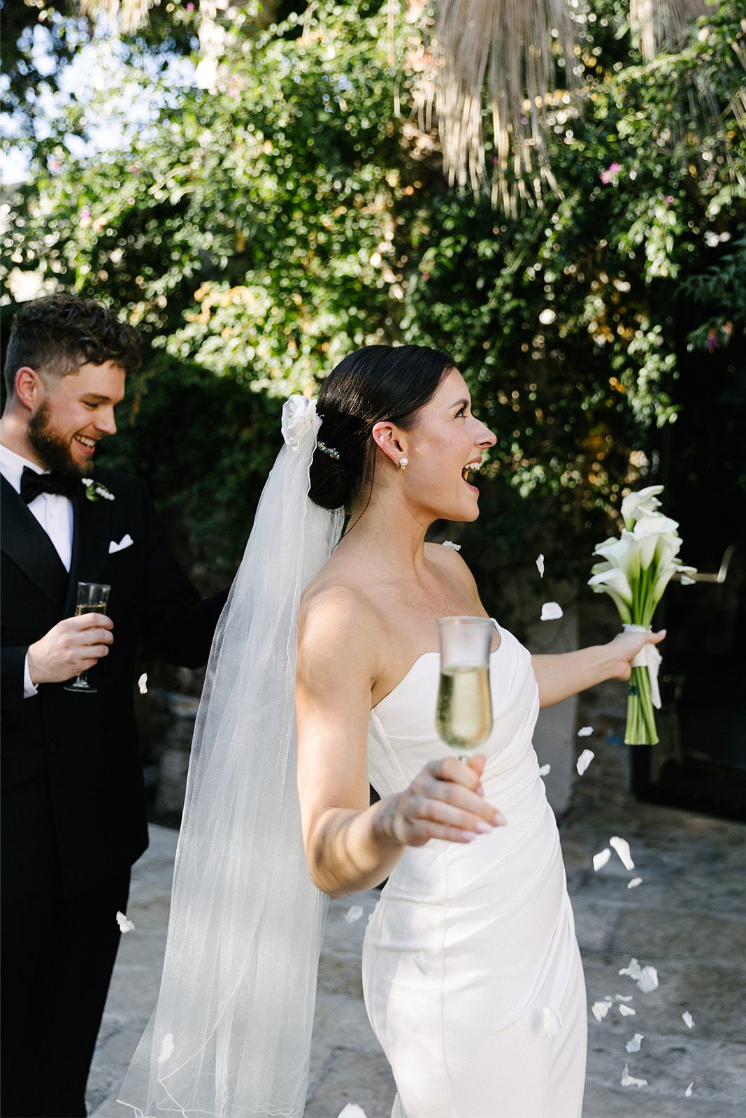 Bride joyfully celebrating with champagne and flowers, groom smiling in background.