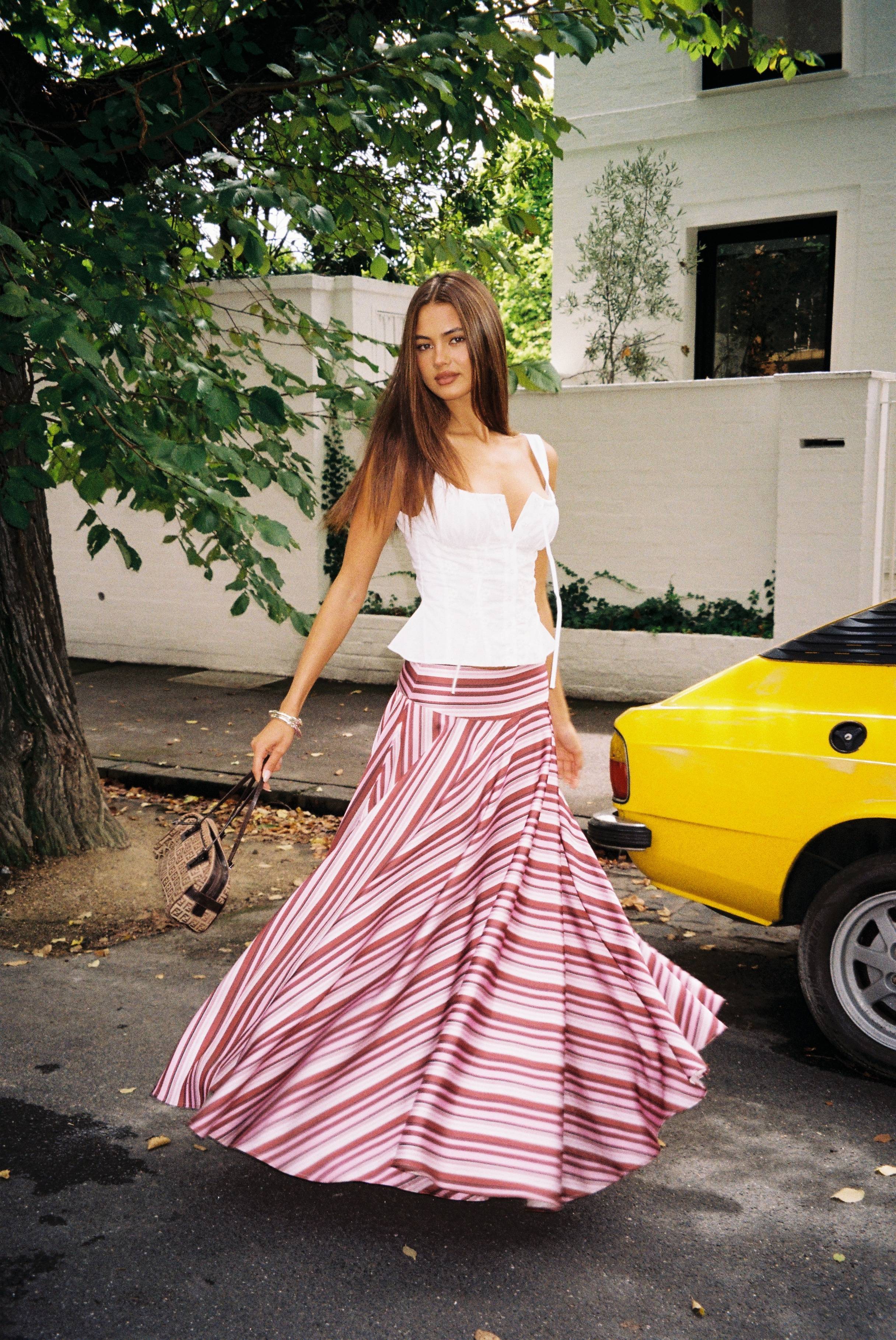 A woman in a white top and striped skirt walks near a yellow car.