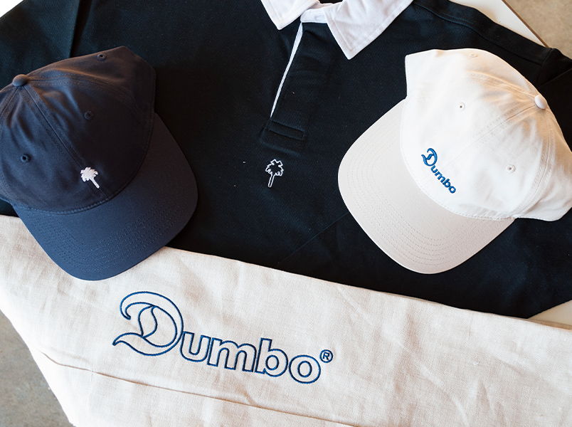 A merch bundle for Dumbo icecream featuring embroidered navy and white cap, rugby jersey, and folded cream apron with a large outline embroidery of the dumbo text