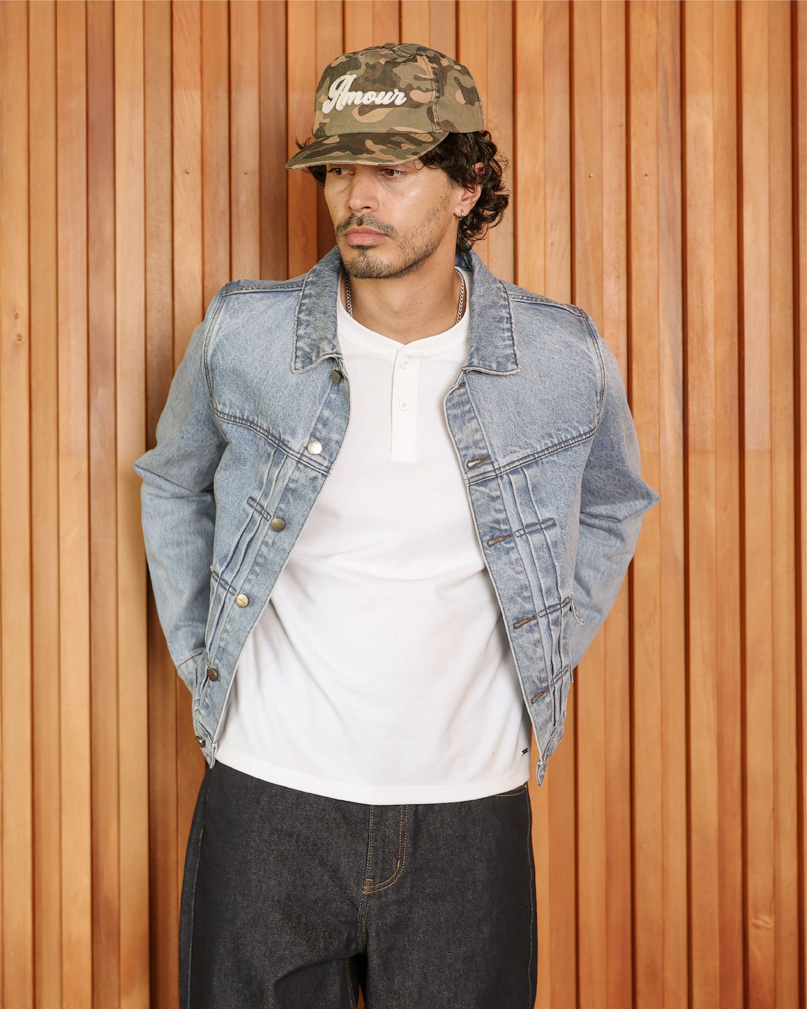 Man wearing a denim jacket and camo cap against a wooden wall.