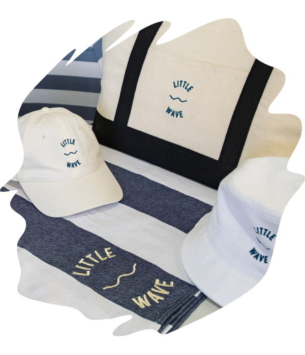 Embroidered merchandise for 'Little Wave' showing the logo in blue thread on bucket hat, cap and bag, and logo in cream on a turkish towel 