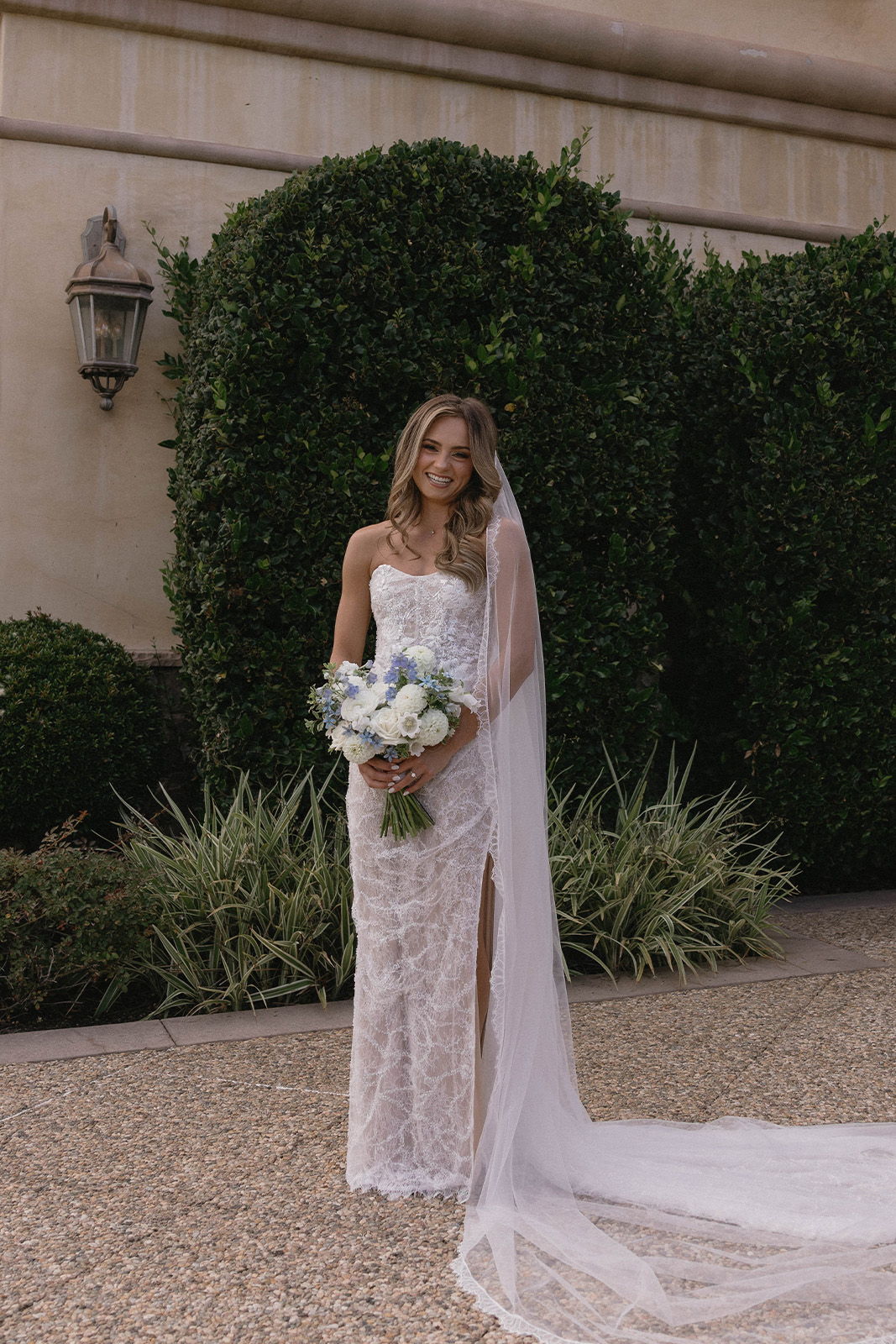 Bride in a lace wedding dress holding a bouquet, standing outdoors.