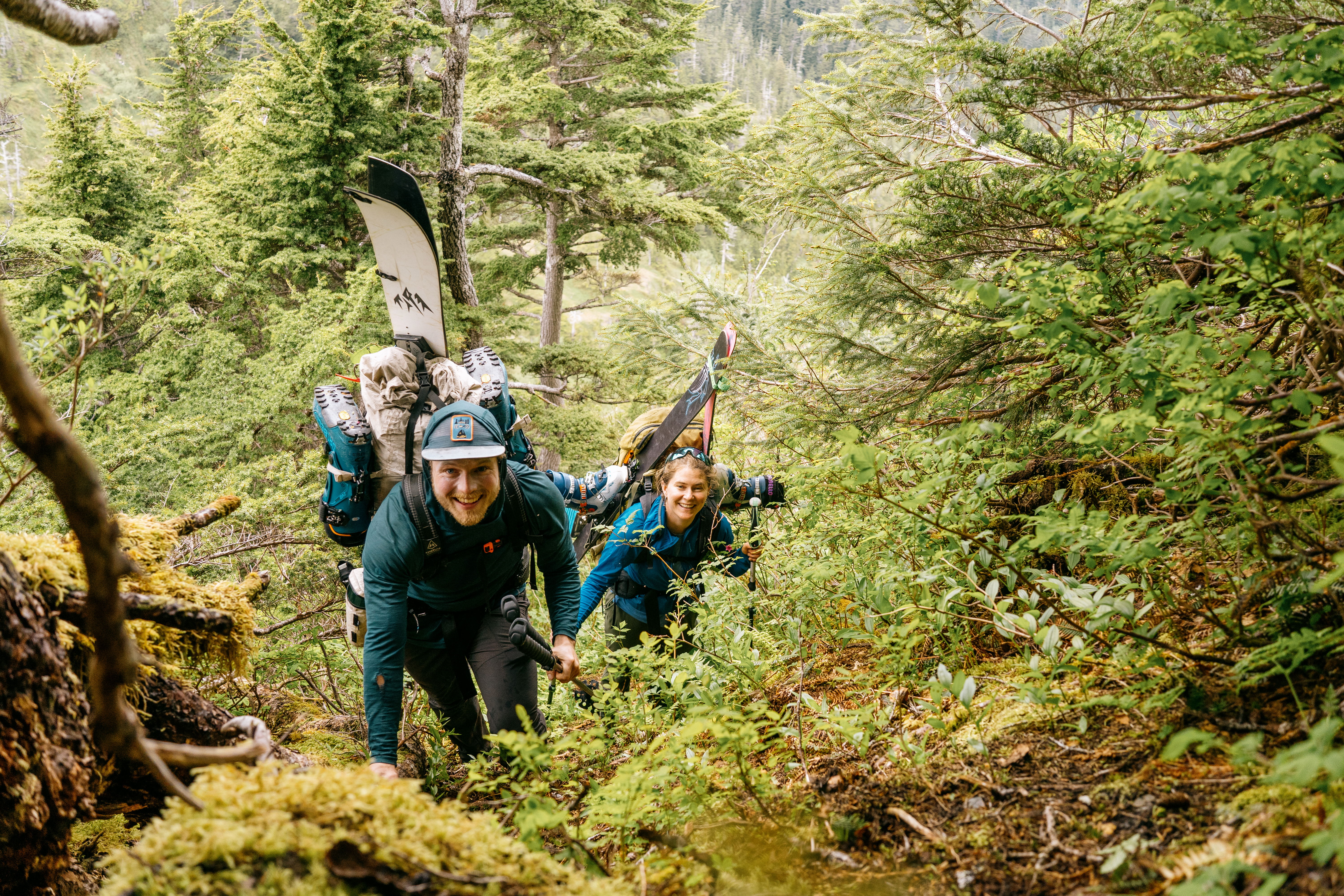 Two splitboarders climbing a forested trail, carrying backpacks and splitboards.