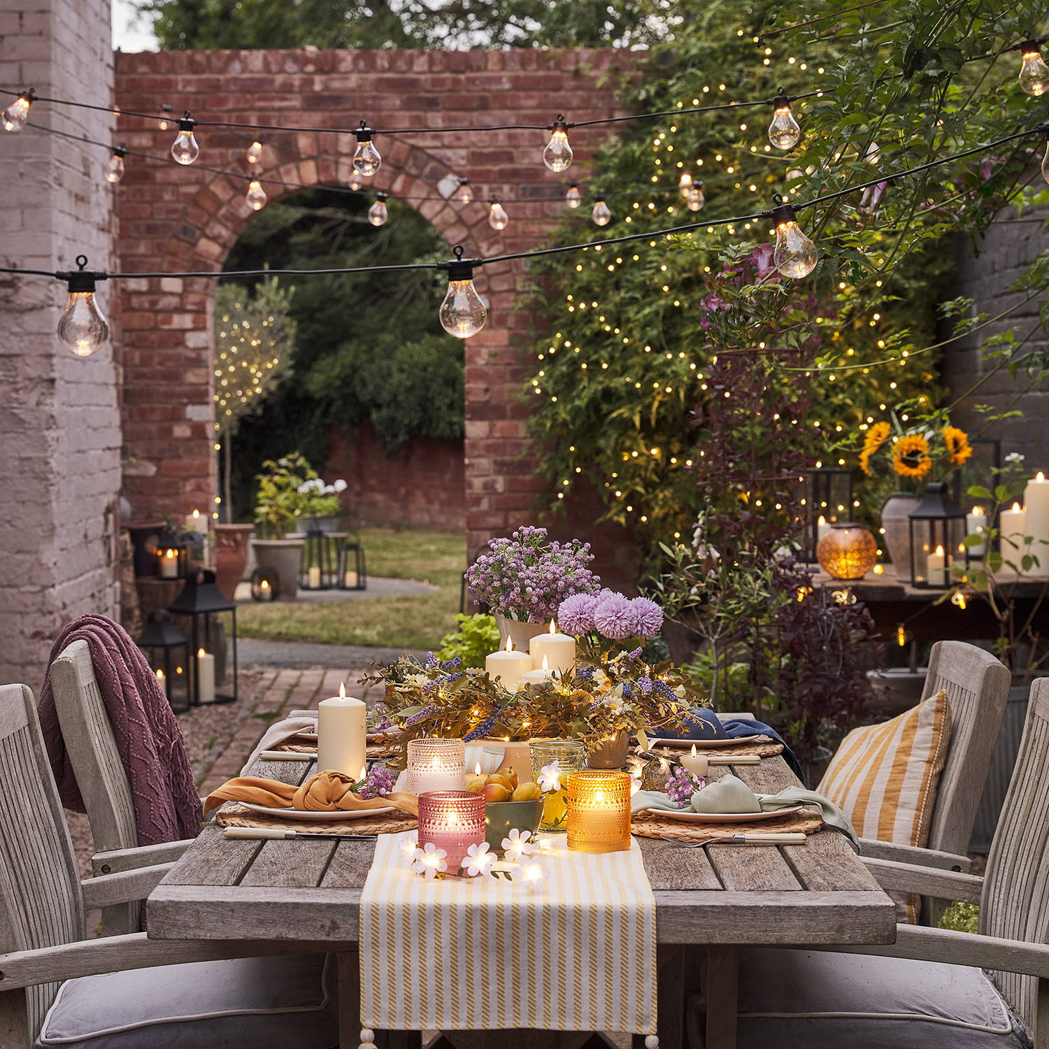 An outdoor alfresco dining setting with festoon lights hanging above the table with lanterns fairy lights and LED candles in the background.