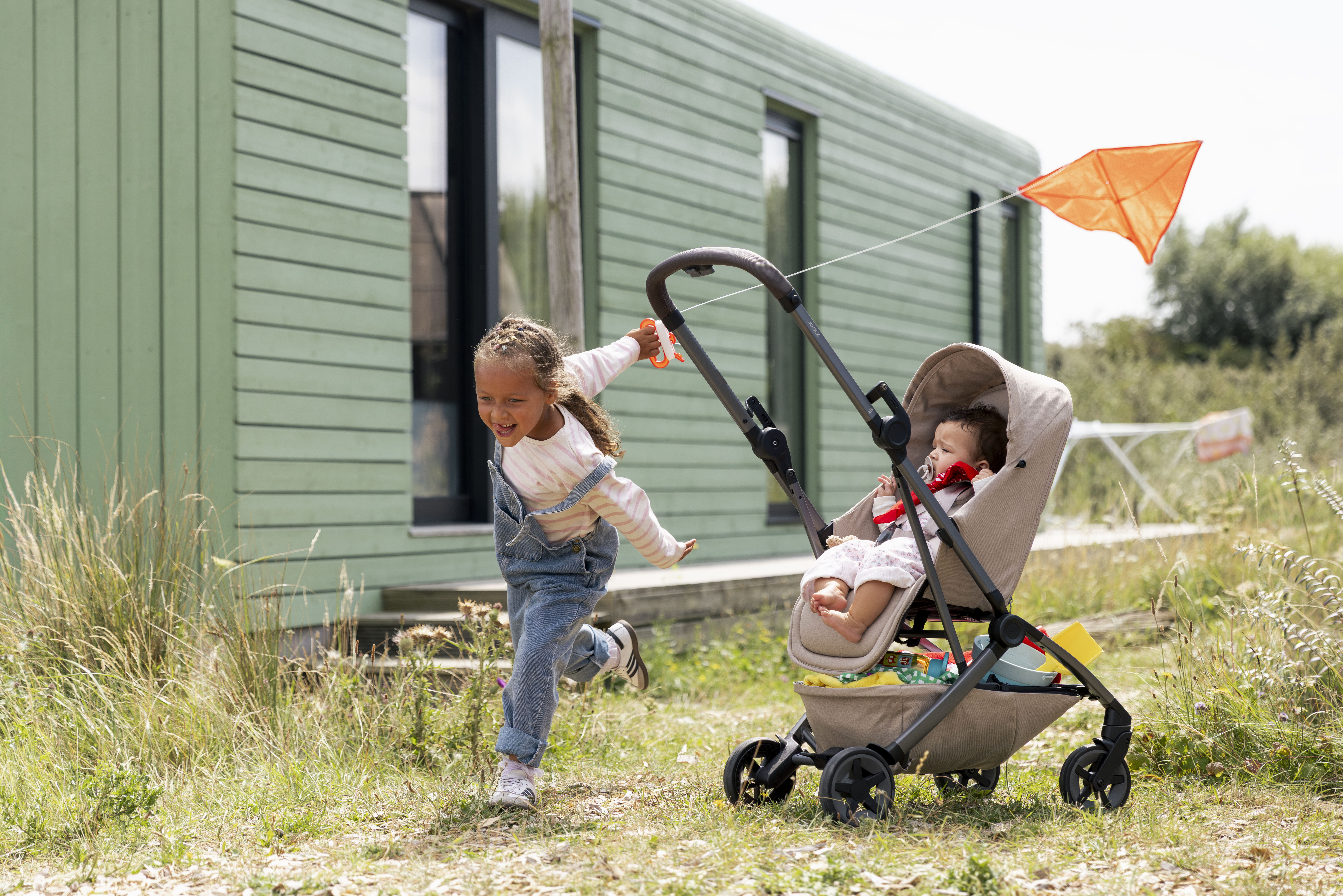 Child playing near a stroller with another child inside, green building in background.