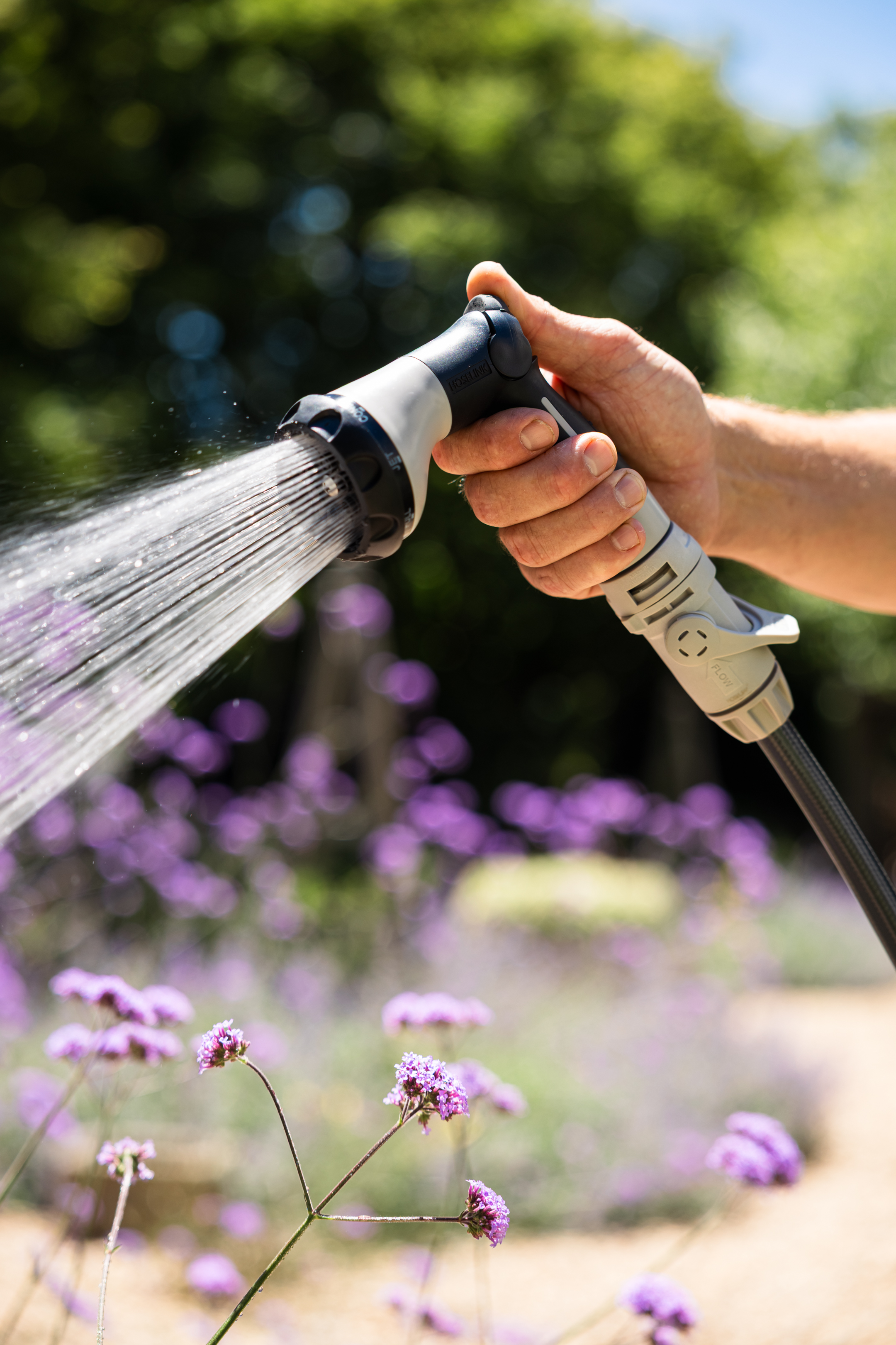 A hand holding a Hoselink 8-pattern flow control sprayer watering purple flowers.