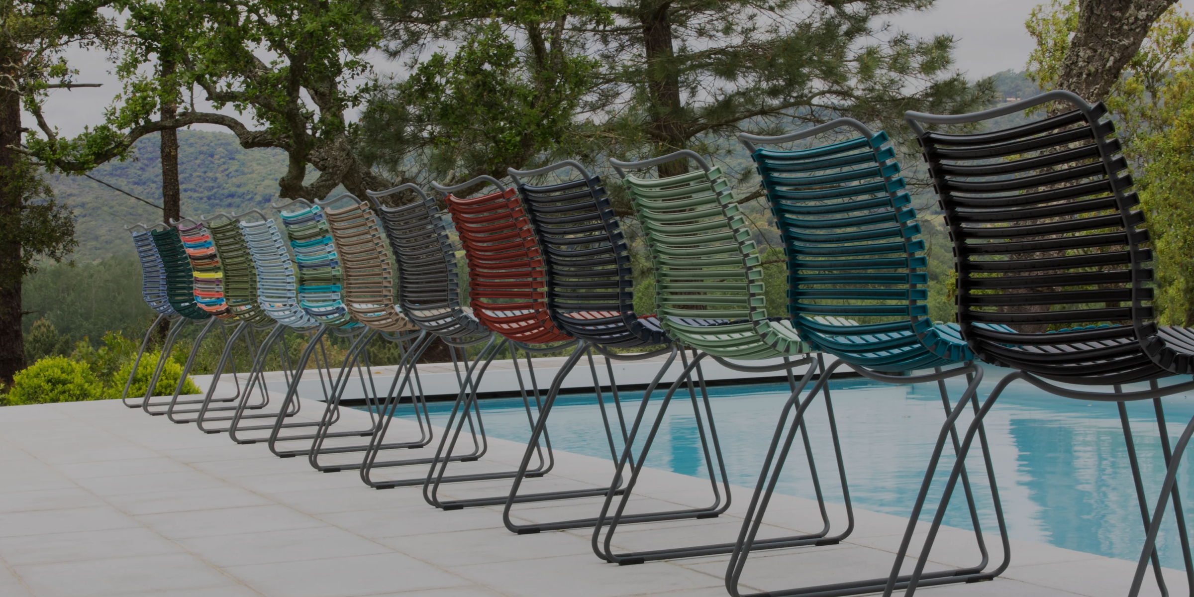 Row of colorful outdoor dining chairs arranged beside a swimming pool with wooded hills in the background.