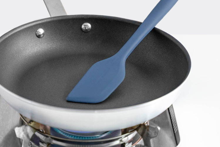 A Blue Misen Mixing Spatula rests in an empty nonstick pan on a gas burner, demonstrating its heat resistance.