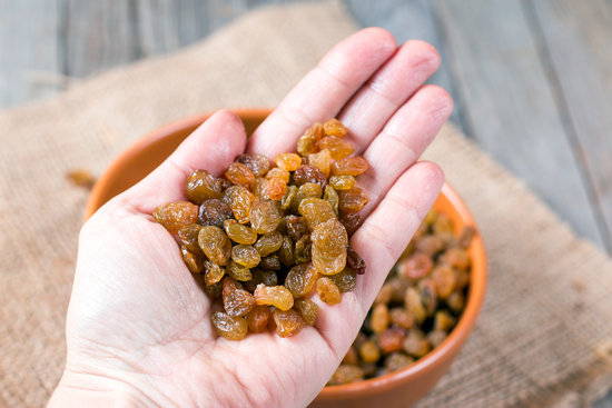 A hand holds a variety of raisins with a bowl of raisins in the background on a table