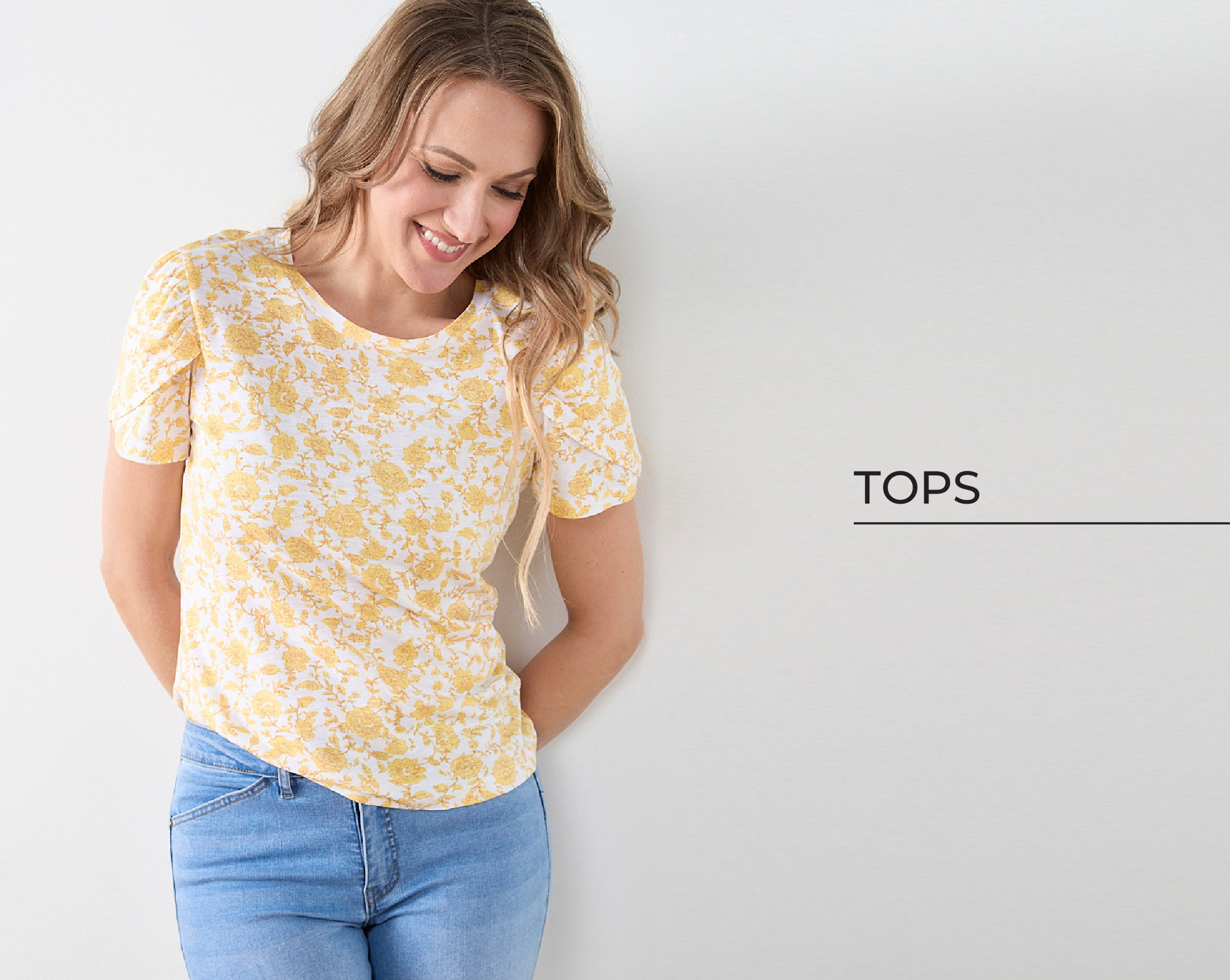 Woman in a yellow floral top against a white wall, smiling.