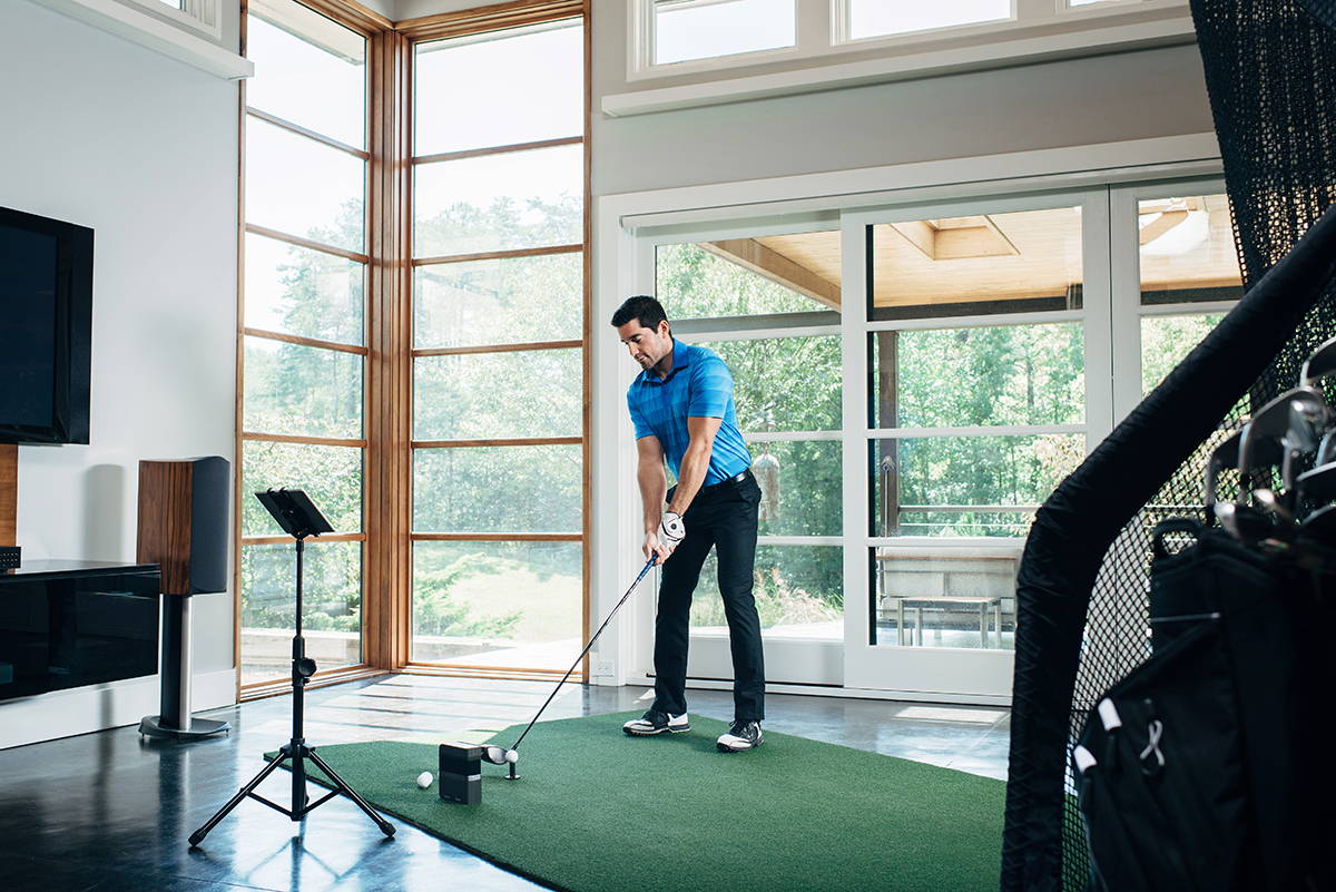 A golfer in his home golf simulator setup with a SkyTrak unit, golf practice net and tablet on a stand