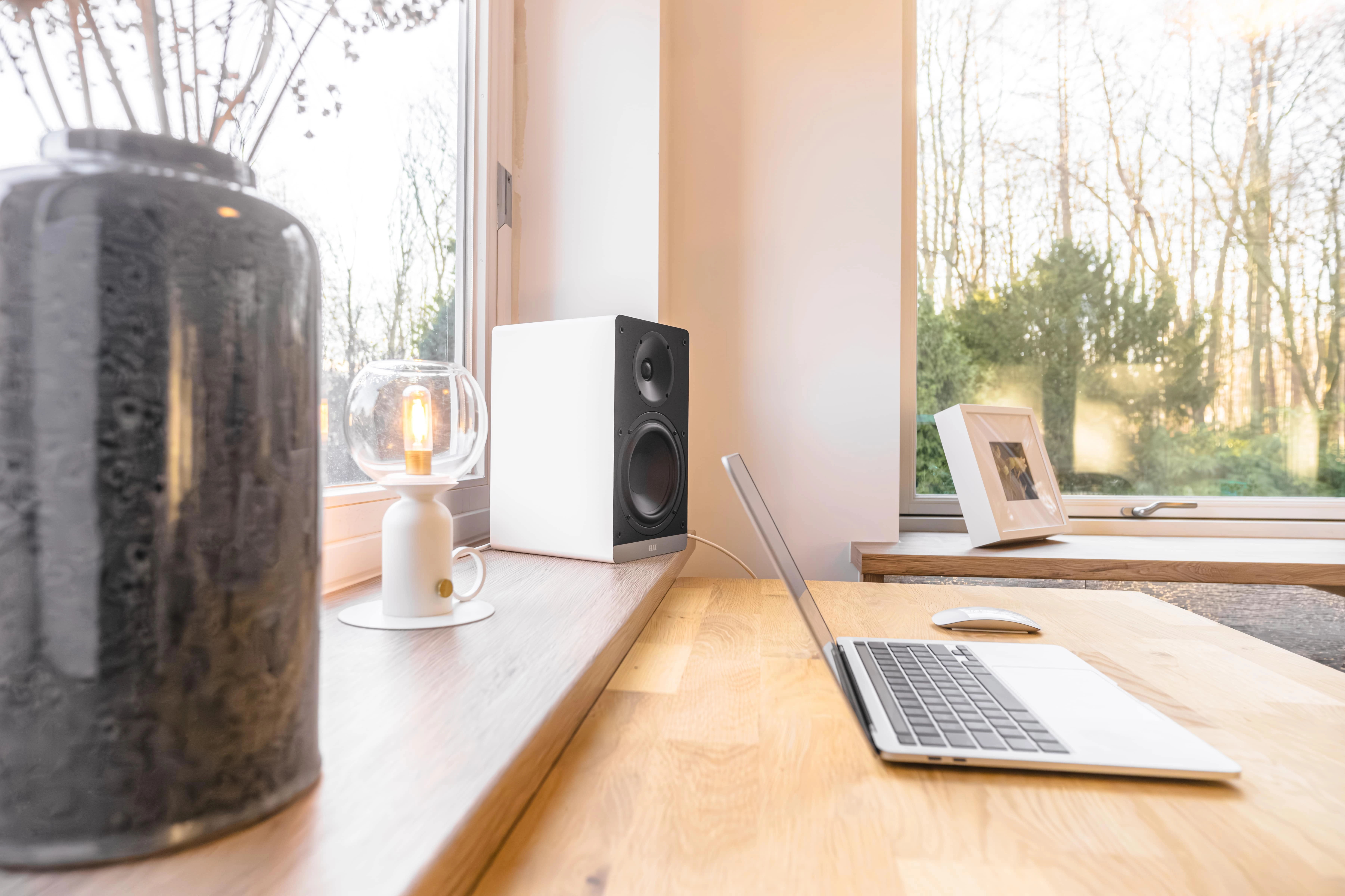 A modern workspace with a laptop, speaker, and decorative vase by a window.