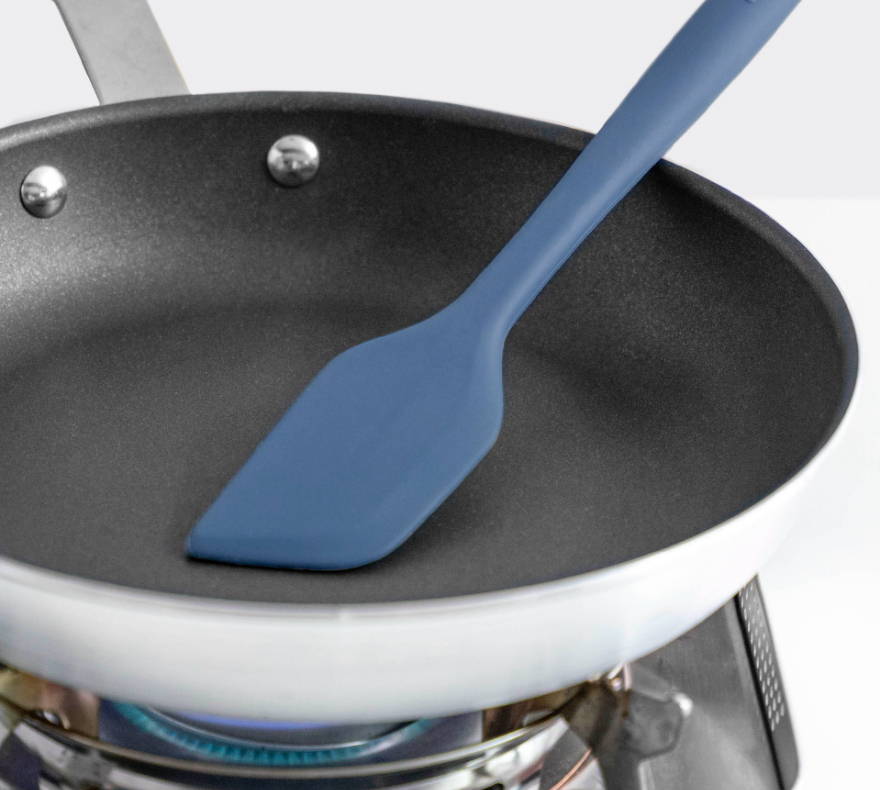 A Blue Misen Mixing Spatula rests in an empty nonstick pan on a gas burner, demonstrating its heat resistance.