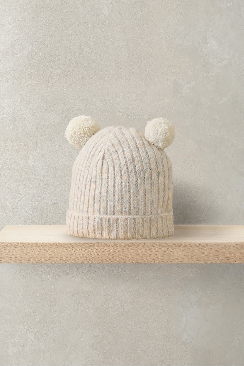 A light-colored knitted hat with pom-poms on top, displayed on a shelf.
