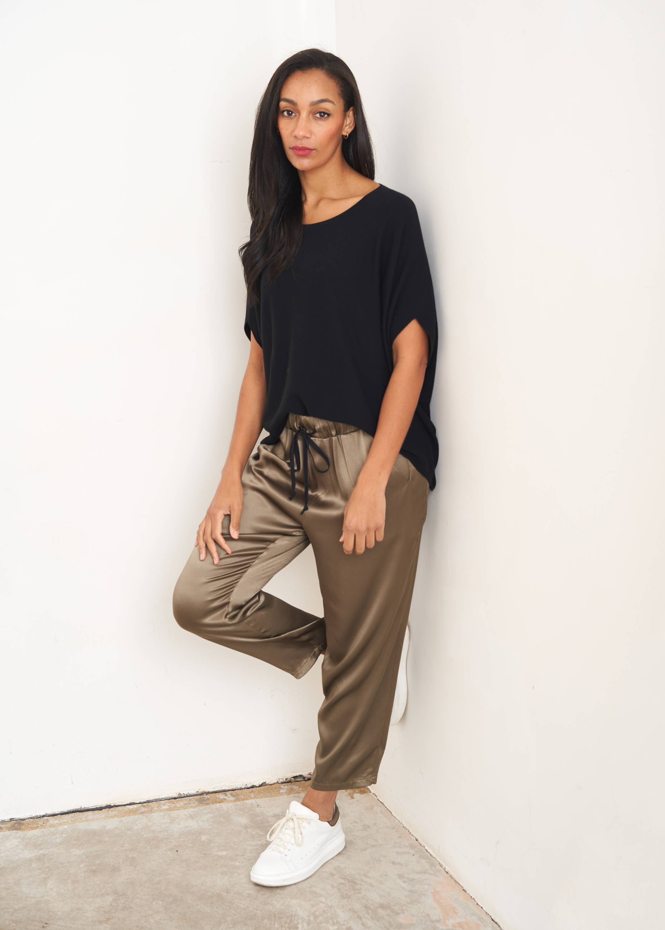 A model wearing a black capped sleeve knitted top with khaki coloured straight leg satin trousers and white trainers