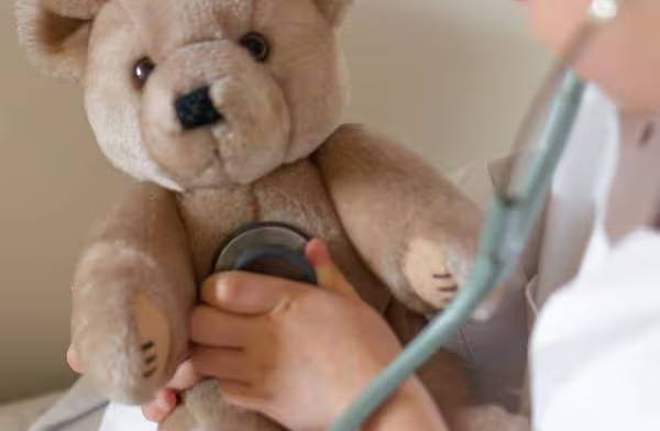  Ein Kind im weißen Kittel spielt Arzt – es verwendet ein Stethoskop, um einen Teddy zu untersuchen