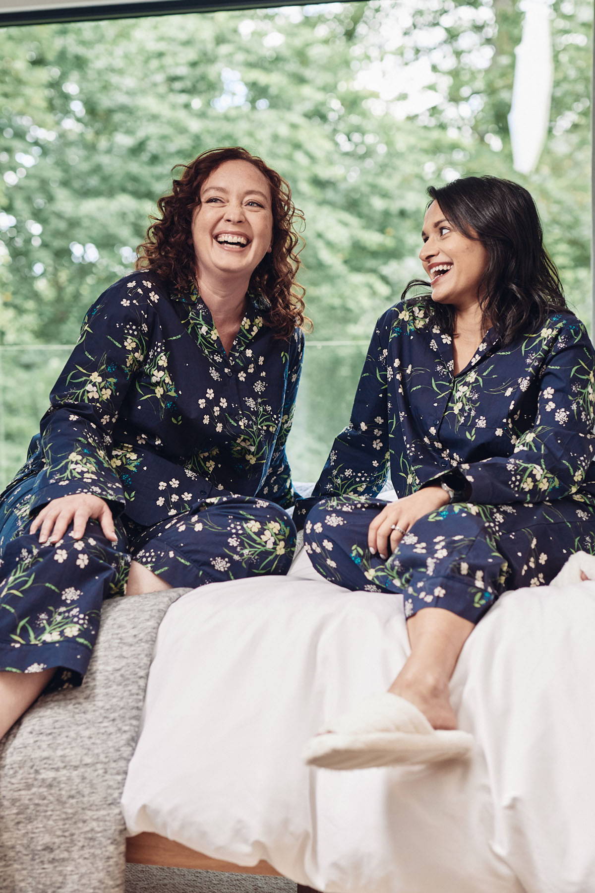 Two women sitting on a bed wearing Rosie Sustainable Pyjamas. The pyjamas are an affordable, sustainable PJ set- in navy with a floral print, made from sustainably sourced cotton.