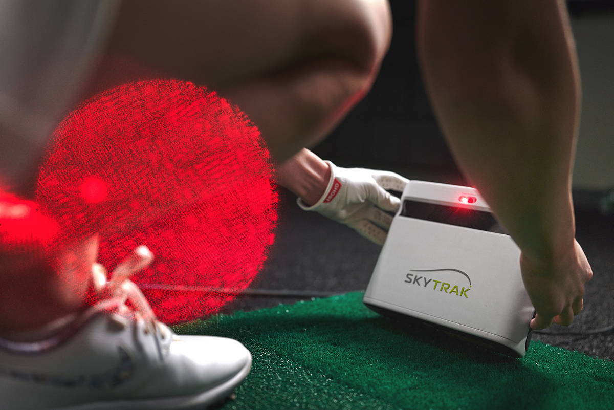 Closeup of a woman squatting to adjust a SkyTrak+ golf launch monitor unit on a golf mat