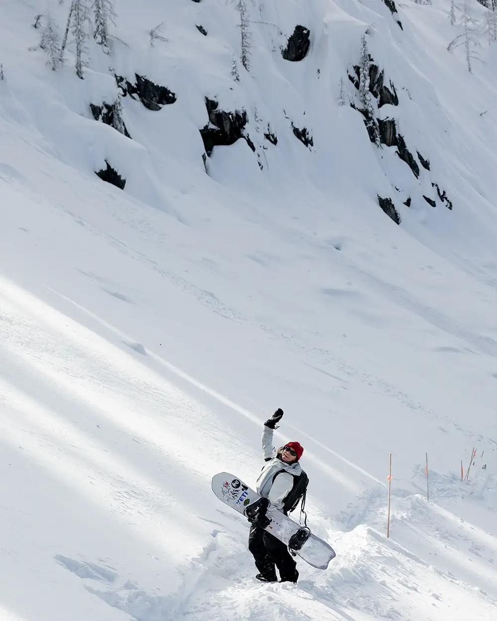 A snowboarder in snowy terrain raises one hand, surrounded by trees and rocks.