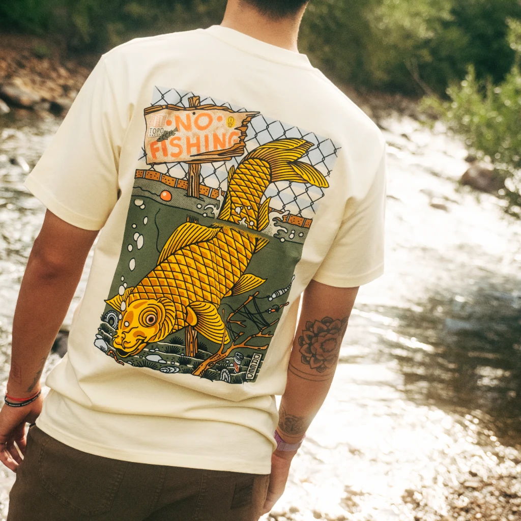 Person wearing a t-shirt with a colorful fish graphic by a river.