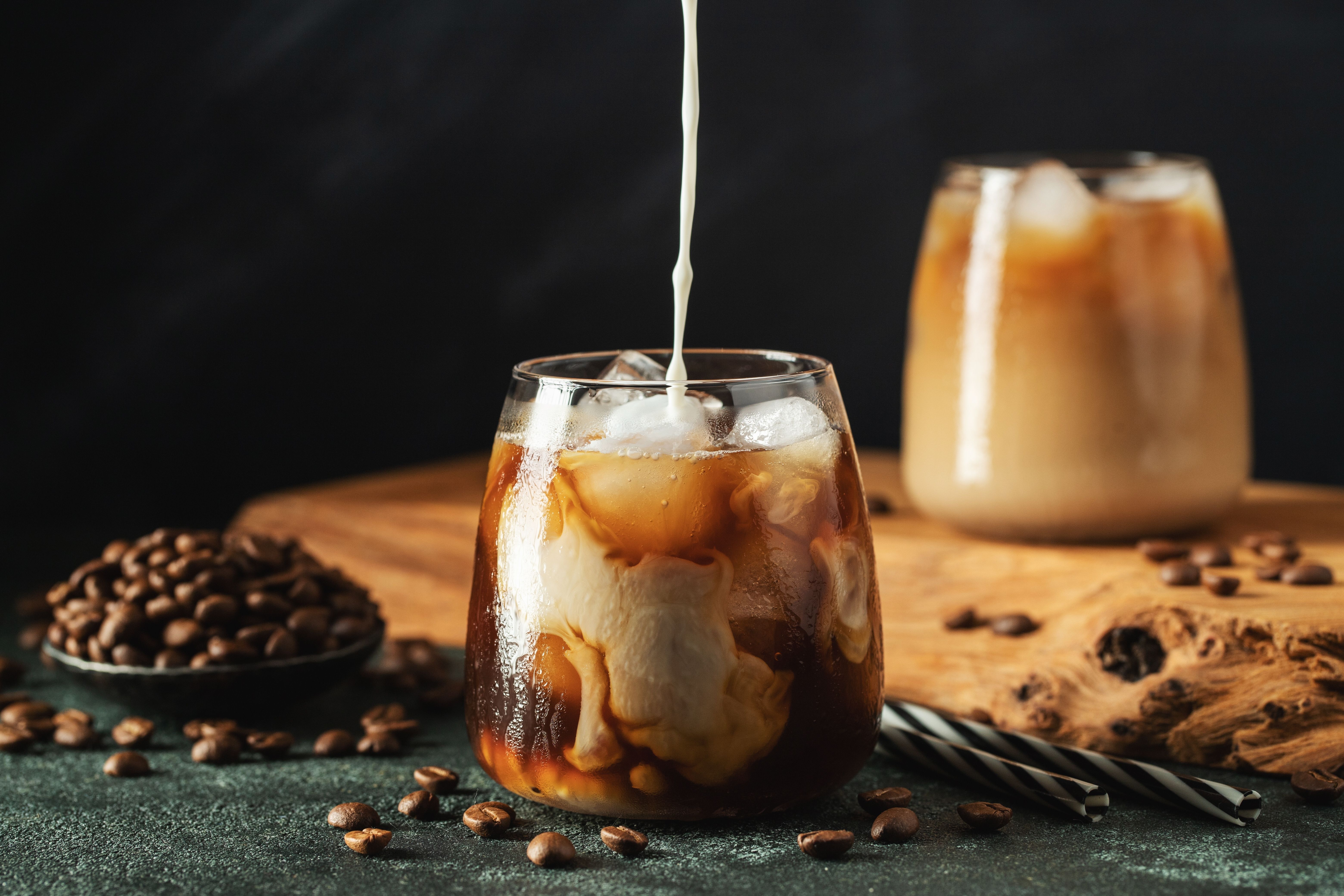 Iced coffee being poured into a glass with coffee beans nearby.