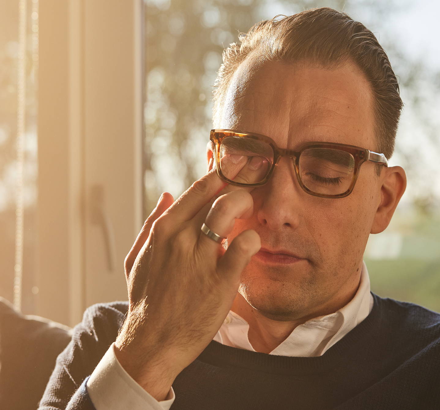 Ein Mann reibt sich ein Auge hinter seiner Brille, während er zu Hause auf seinem Sofa sitzt – Eine Hausstaubmilbenallergie kann zu roten, juckenden und tränenden Augen führen.