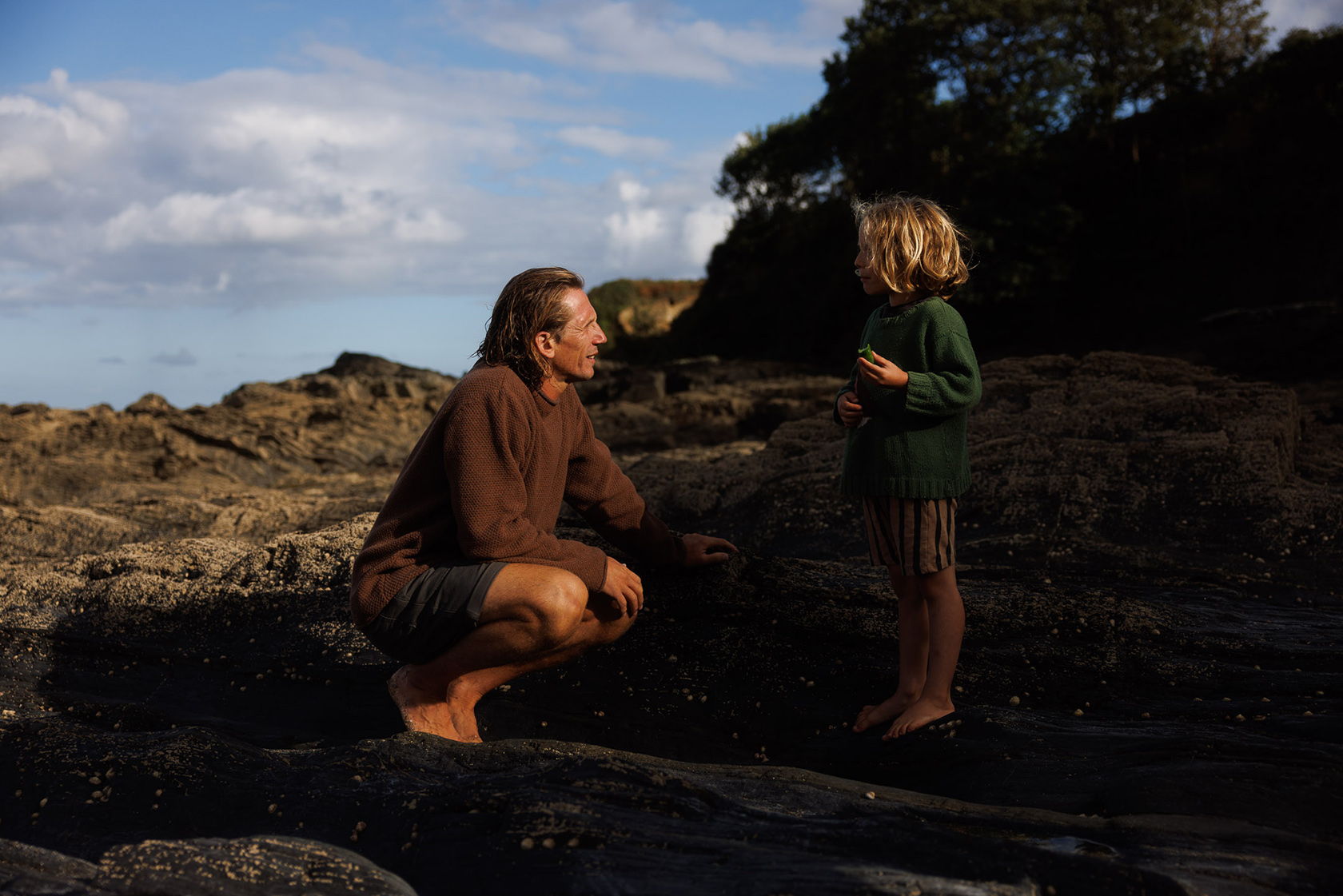 A man and a child interacting on rocky shoreline under a cloudy sky.