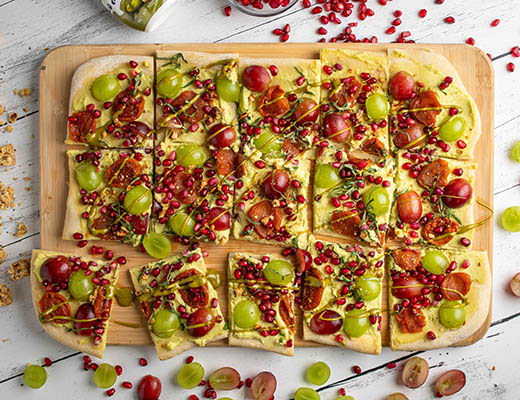 A dessert pizza topped with grapes, pomegranate seeds, and nuts on a wooden board.