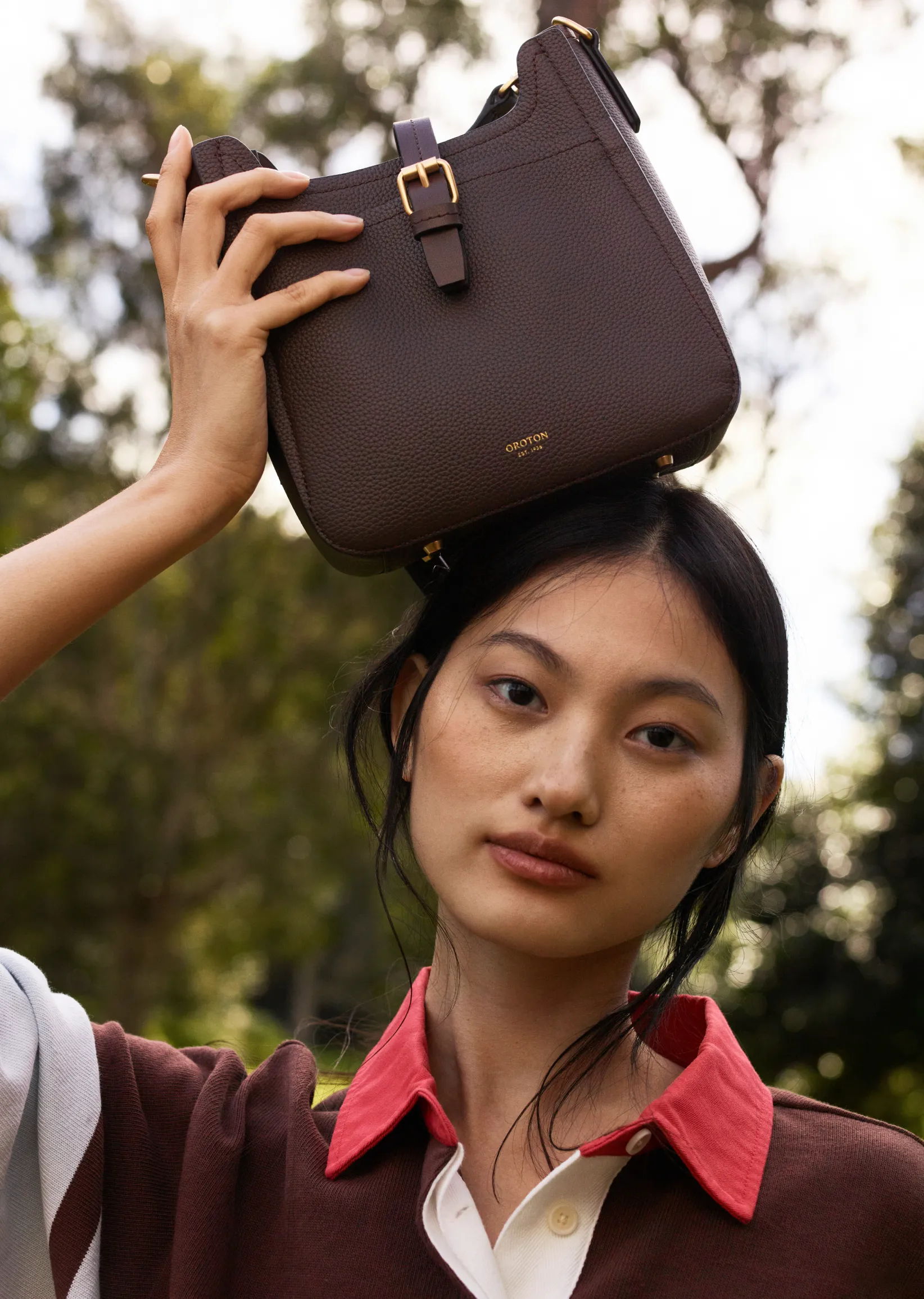 A woman holding a brown handbag above her head outdoors.