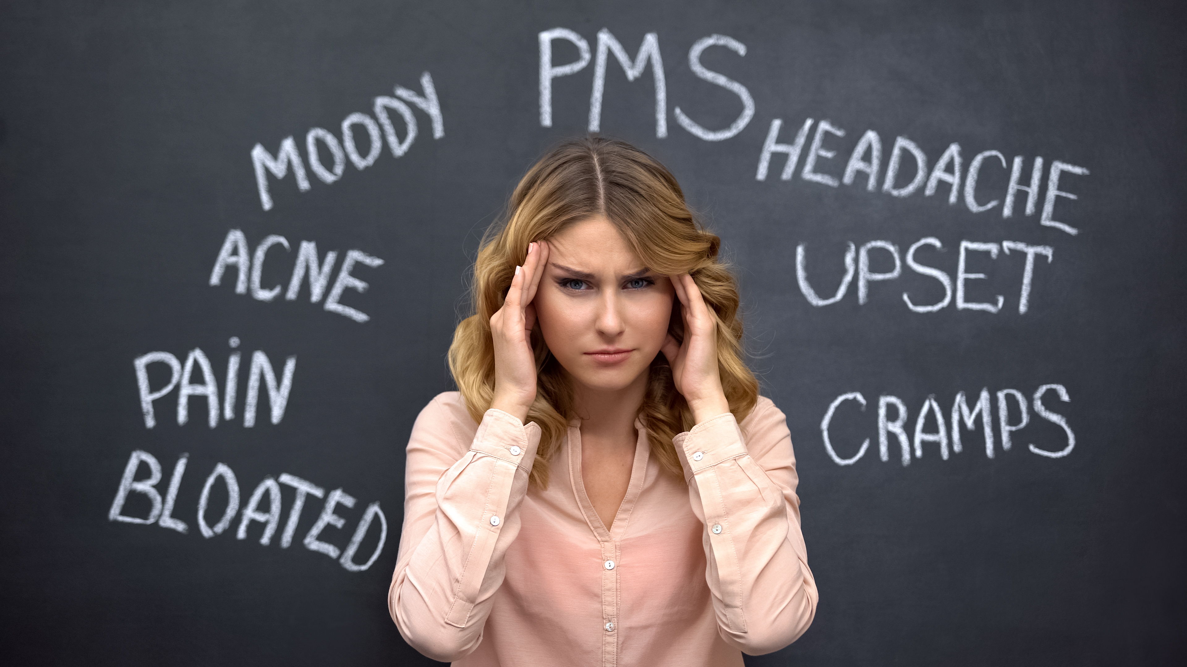 Woman with headache surrounded by PMS-related words on a chalkboard.