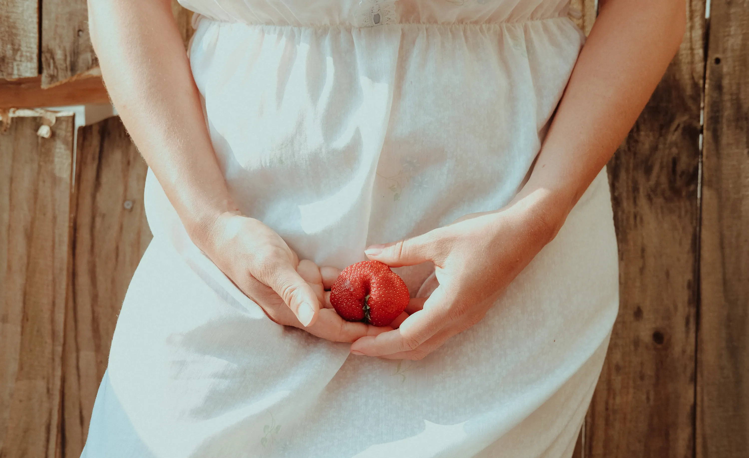  A woman in a white dress stands in front of a wooden fence holding a piece of fruit in front of her hips.