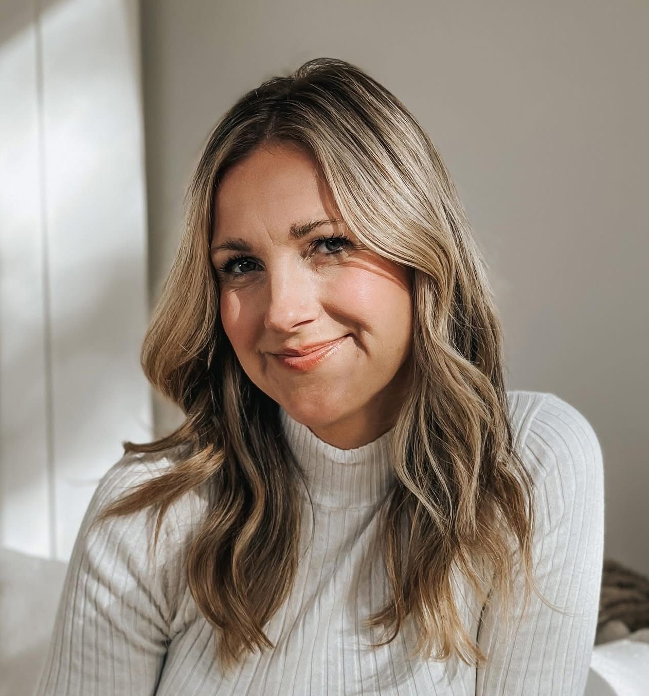 Smiling woman with wavy hair wearing a white sweater in soft lighting.
