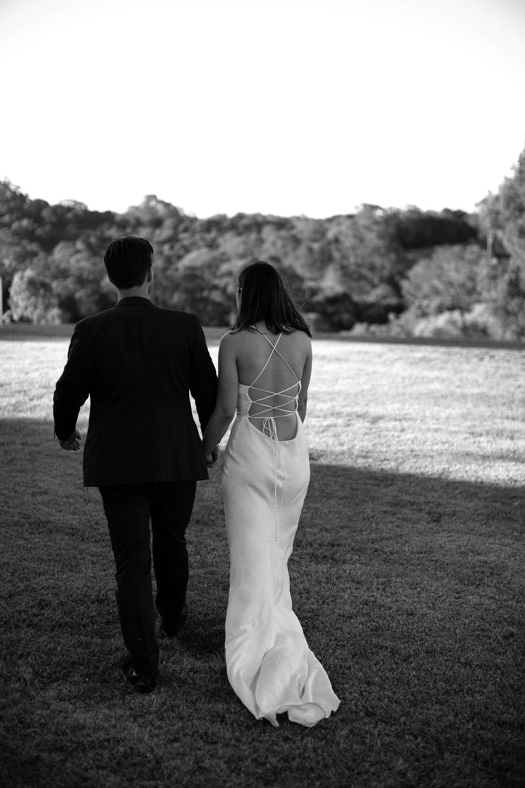 Novia con un vestido de seda atado a la espalda caminando en el campo de hierba con su novio