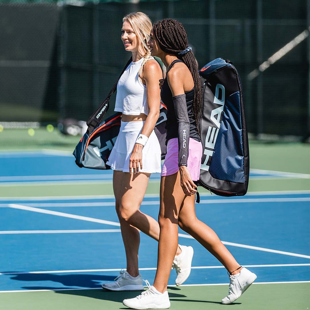Two tennis players cross a tennis court carrying their HEAD™ gear, including smart compression sleeves on their arms.