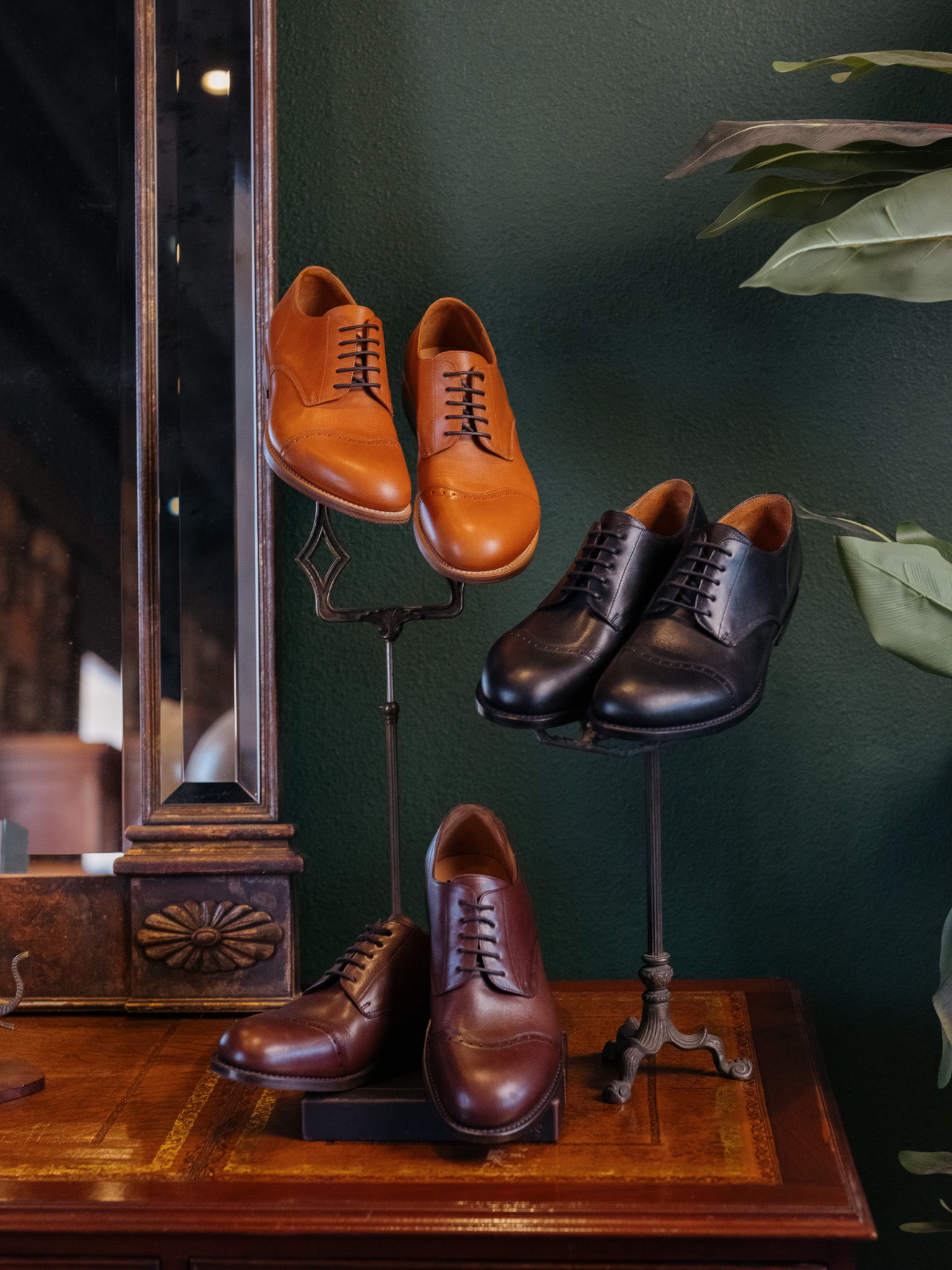 Four pairs of dress shoes displayed on stands against a green wall.