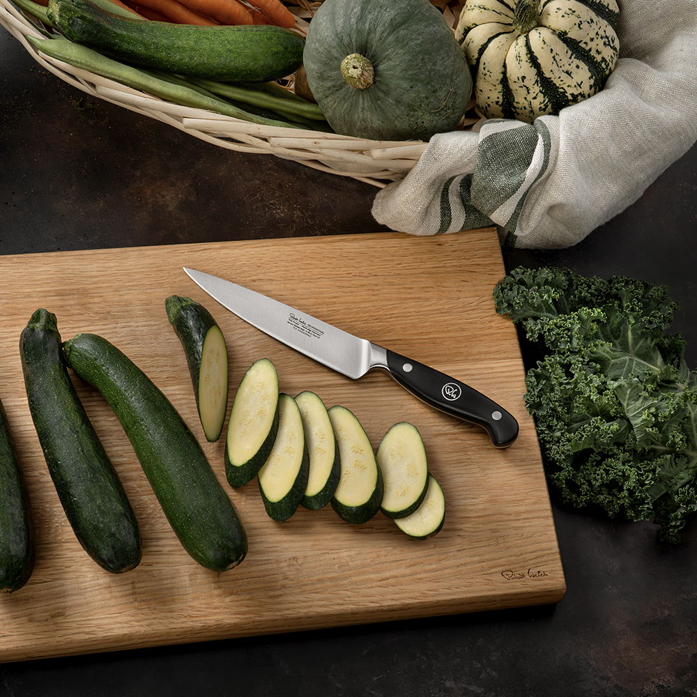 Sliced courgettes on a cutting board with the 14cm Professional Kitchen Knife and vegetables in a basket.