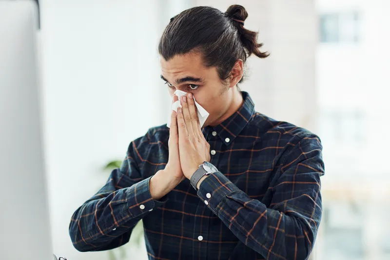 Man met zwart geruit overhemd met zwart haar in een knotje, snuit zijn neus in een tissue. Een loopneus kan een symptoom zijn van allergie.