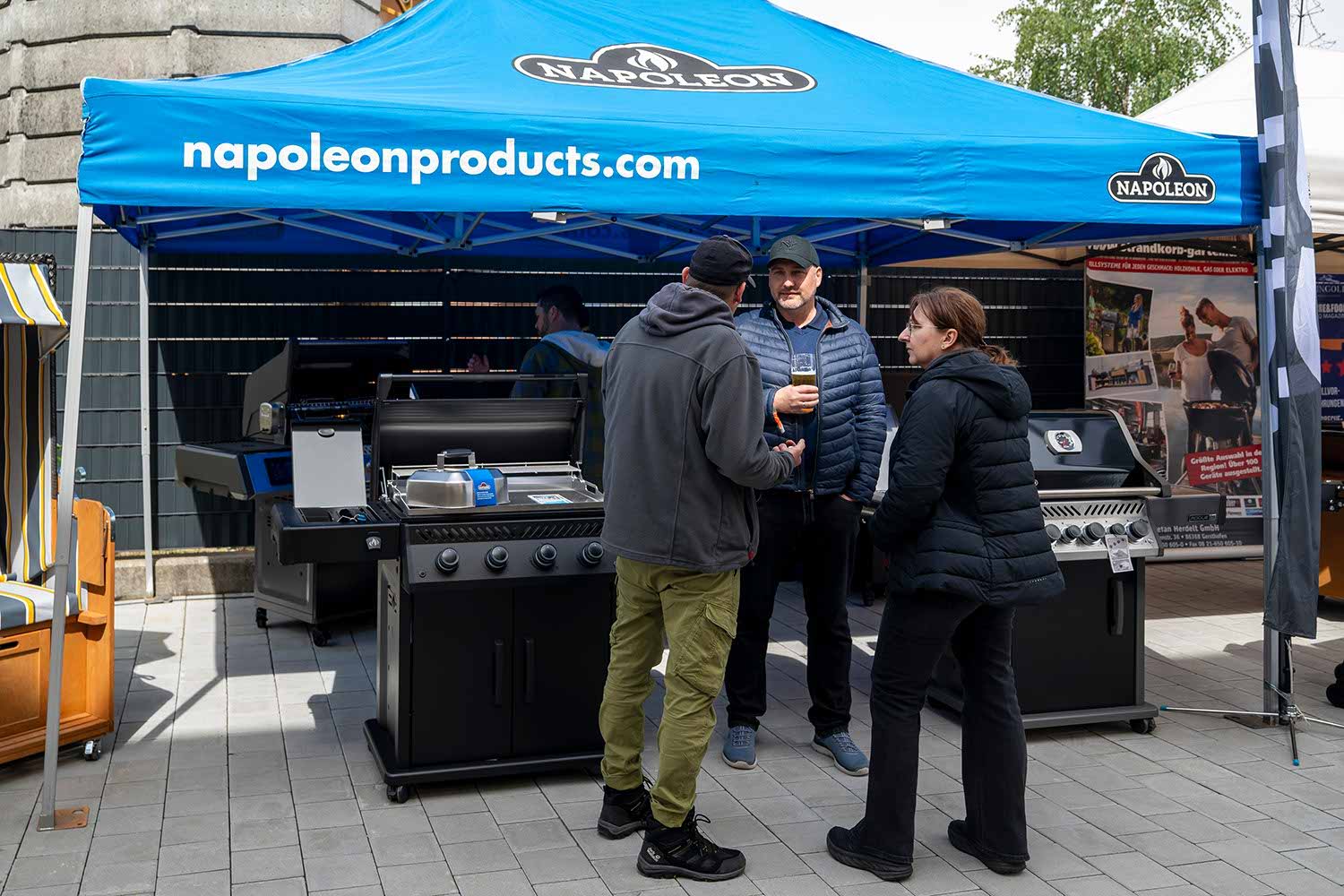 People discussing grills under a blue canopy at an outdoor event.