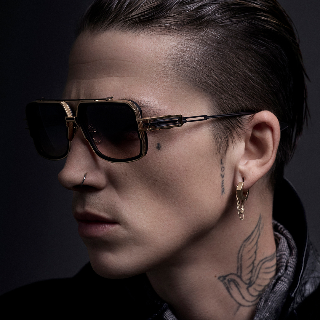 Profile of a person wearing sunglasses and a tattoo, against a dark background.