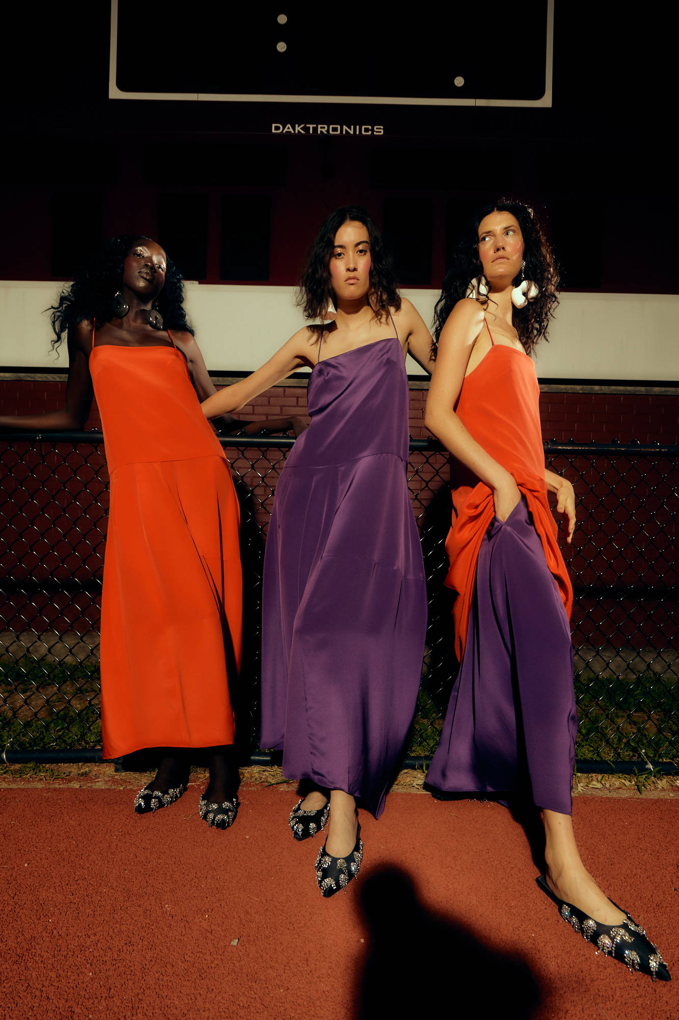 Three models in elegant dresses pose on a sports court at night.