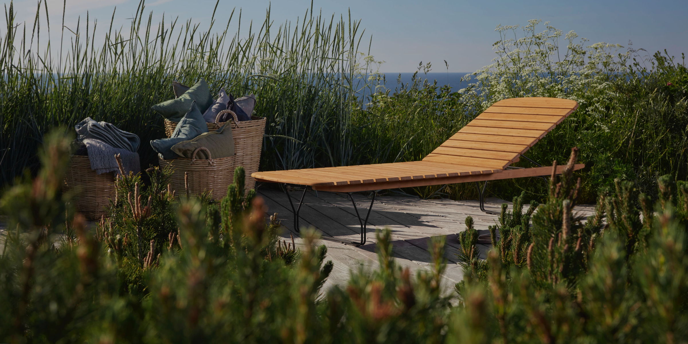 A teak chaise lounge on a beachside deck surrounded by green plants