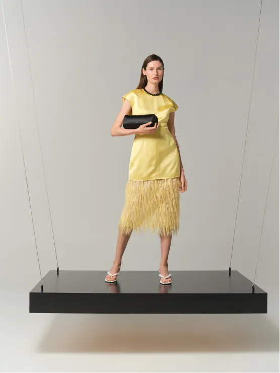 Model in yellow dress with fringe, holding a black clutch on a suspended platform.