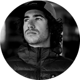 Man with curly hair wearing a hat and jacket, looking thoughtfully upwards.