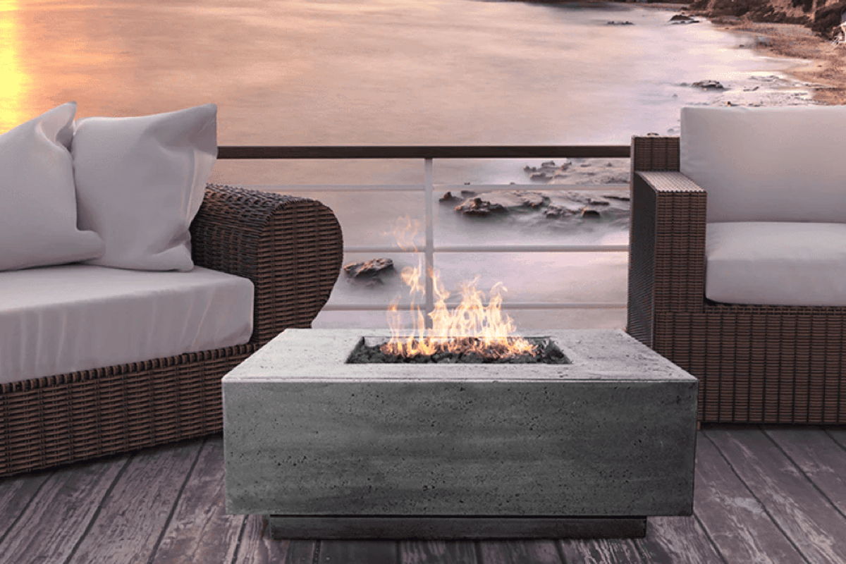 Concrete rectangular fire pit with flames on a deck between two wicker lounge chairs, overlooking the ocean at sunset.