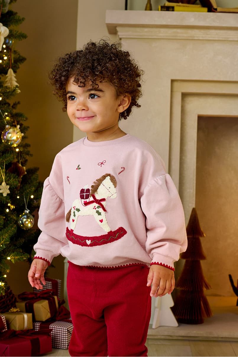 Child in festive sweater, smiling near a decorated Christmas tree and fireplace.