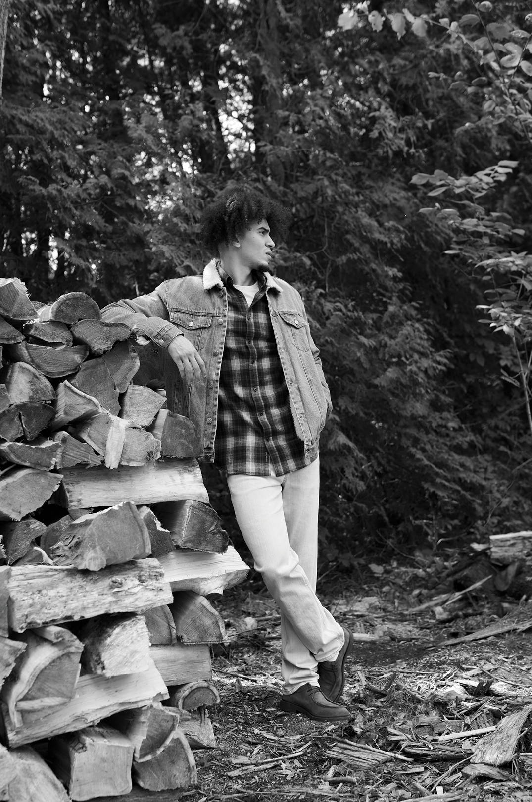 Tall man leaning against a pile of wood in a plaid shirt and denim jacket