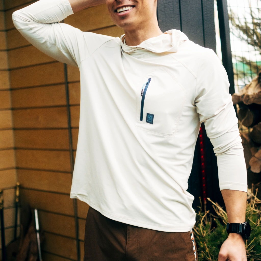Smiling person wearing a light hoodie, posing outdoors near a wooden wall.