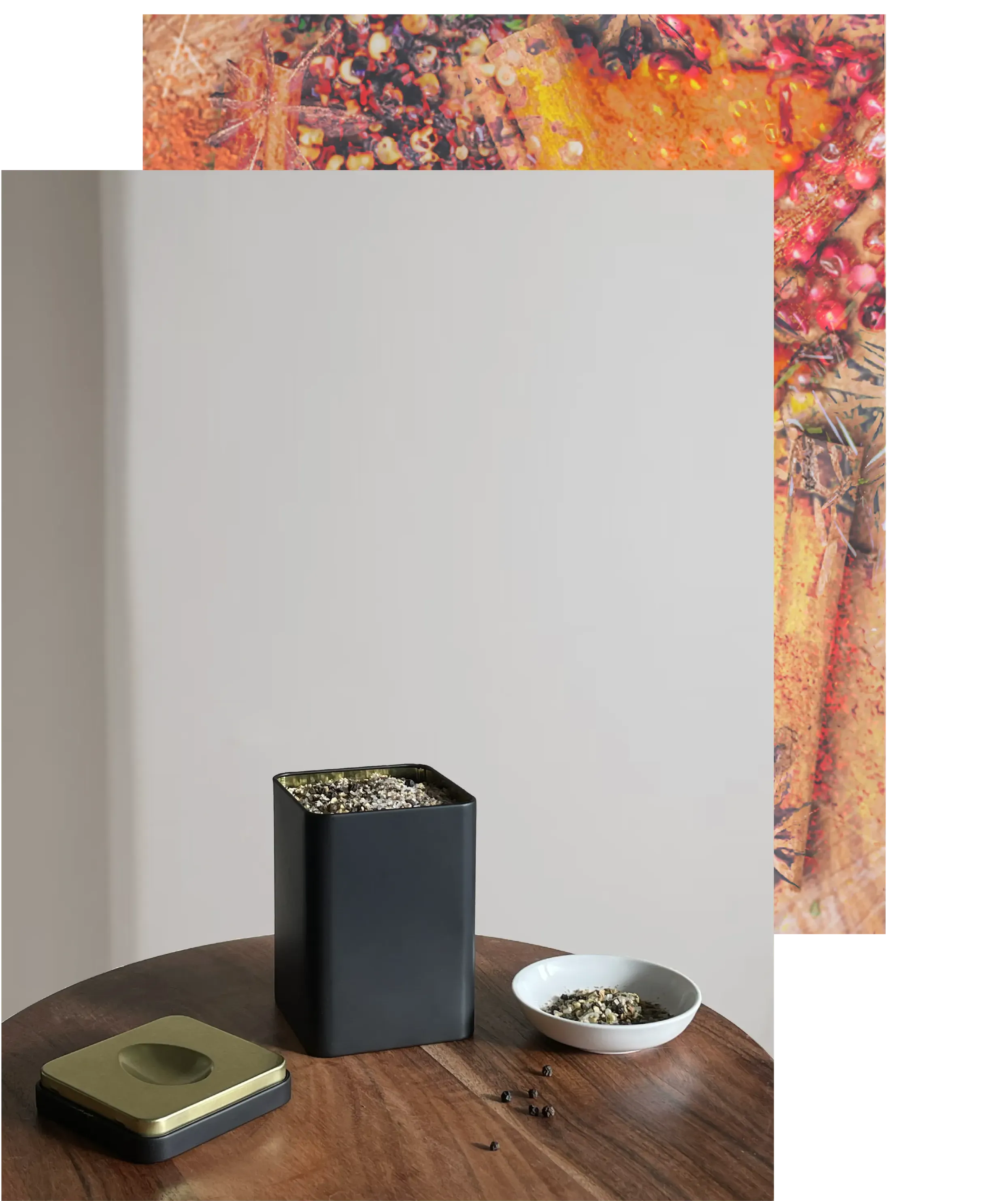 loose leaf tea packaging with holiday spice mix in tin with bowl on the right side on a wooden table the lid of the tin on the left side  loose pepper corn on the table. An image of a stylized mix of holiday spices is in the background.