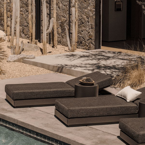Low-profile outdoor chaise lounges with dark performance cushions beside a pool in a modern desert backyard with cactus and stone walls.