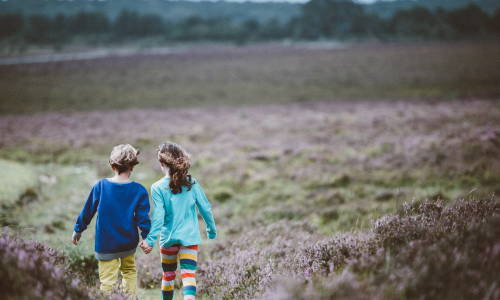 Zwei Kinder, ein Mädchen und ein Junge, marschieren Hand in Hand durch die Heide. Sie scheinen ein Abenteuer zu erleben